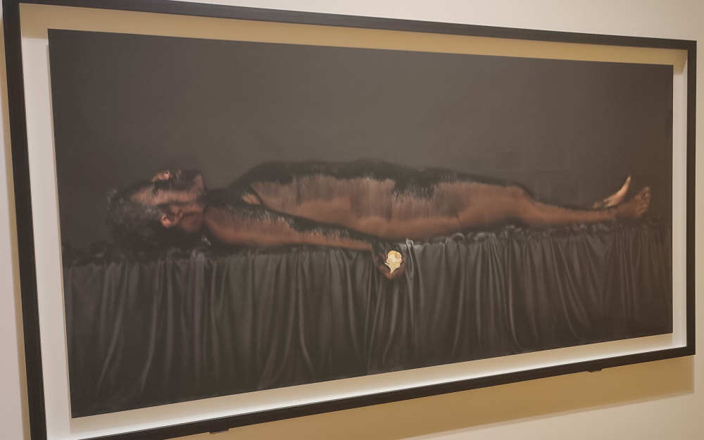 A painting of a black woman covered in coal laying on a cloth-covered black table, as if deceased