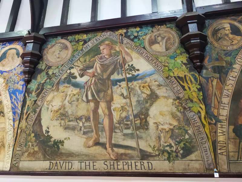 A pre-Raphaelite painting on a wooden panel adorns the walls of the previously depicted church hall. On it, a golden-haired boy in a tunic holding a staff, David the Shepherd, stands proud, herding his flock of sheep as mountains recede in the background. He is depicted within a triangular frame, the top two edges inscribed with Biblical verse, the bottom edge blaring, in all capital letters, "David. The. Shepherd." Around the frame, two more portraits are inscribed in smaller circles, while the rest is painted with bright green leaves and vines.