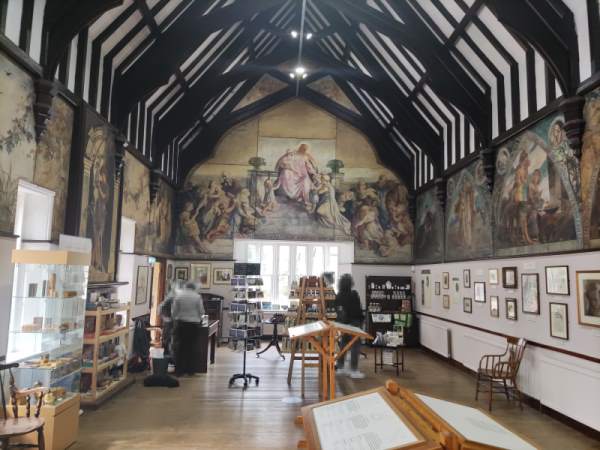 The inside of a small church hall, the upper heights of its walls covered in glorious paintings of Biblical scenes, the lower halves painted white and covered in smaller, framed paintings. The floor is riddled with chairs, information stands, cabinets, and other such auxiliaries. In the back of the shot, two people browse the store shelves, while two others work at the reception desk.