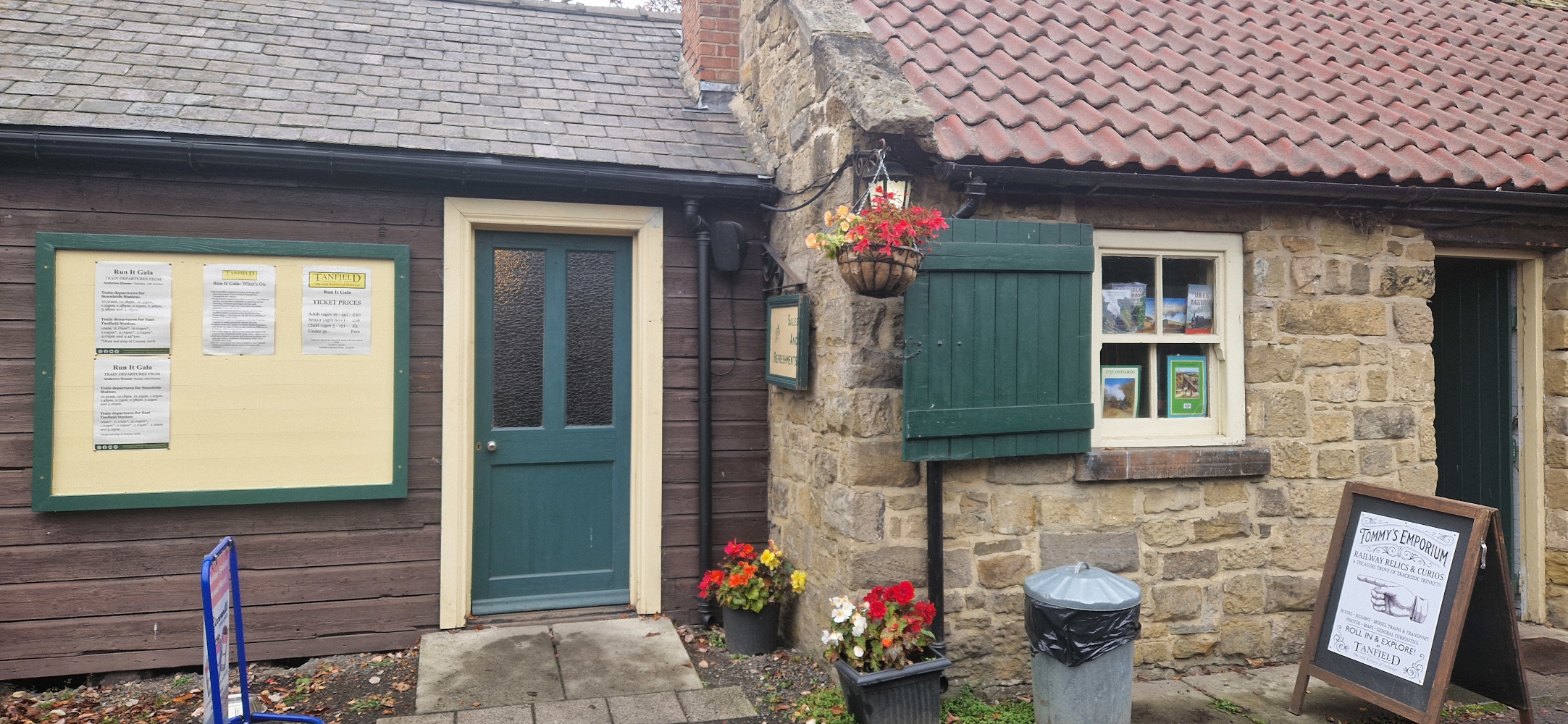 Exterior view of a traditional stone and wooden building at Andrews House station, featuring a green wooden door, window with shutters, noticeboard, hanging flower baskets, and an A-frame sign for "Tommy's Emporium".