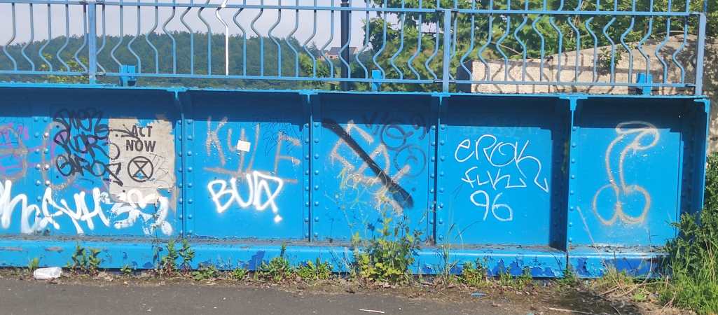 Graffiti op een blauwe brugmuur. Van links naar rechts: Een XR poster met 'Act Now', 'Kyle', 'EDL' (doorgestreept), 'Erok', 'FLK', en een lul en kloten.
