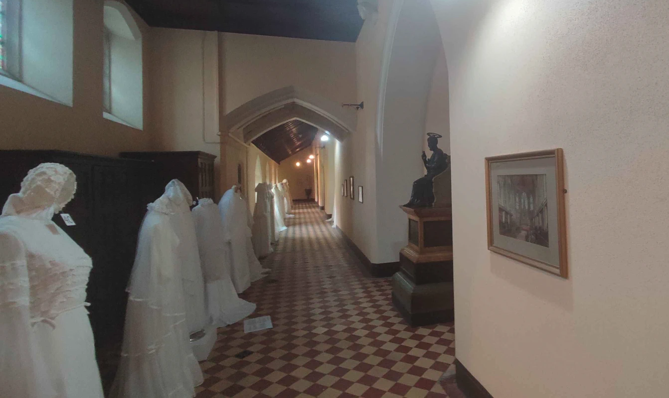 A dimly-lit photo of church corridors with vaulted arches, the plain white walls lined with pictures on one side and dresses on the other. A brass statue of a saint sits in a niche off to the right, and the floor is decorated with a checkerboard of worn white and red tiles.