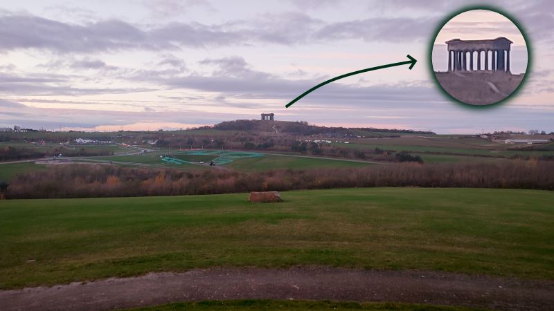 A panoramic view of a sprawling country park, with some noticeable barriers put up for a race. In the distance, upon a hill, lies a building rather resembling an old Greek temple.