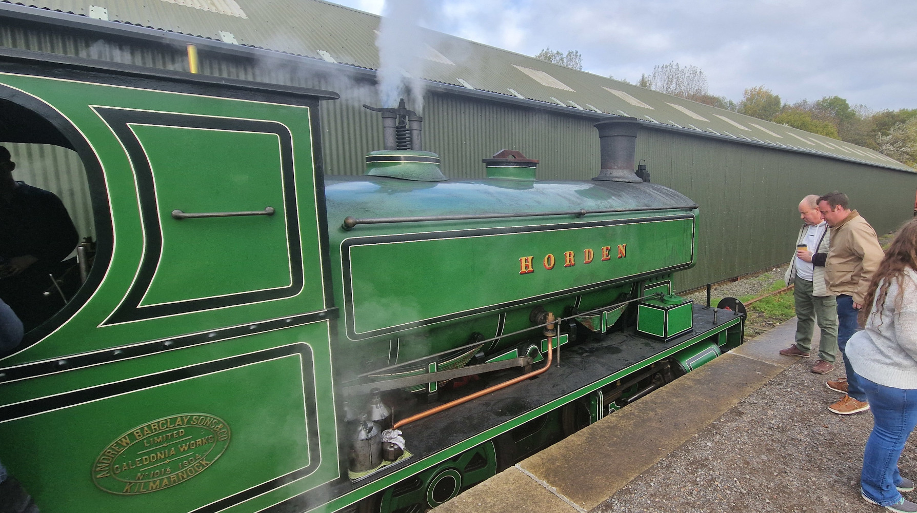 A puffing green steam locomotive labelled “Horden” stands in the bay platform of a small railway station as two older gentlemen watch on