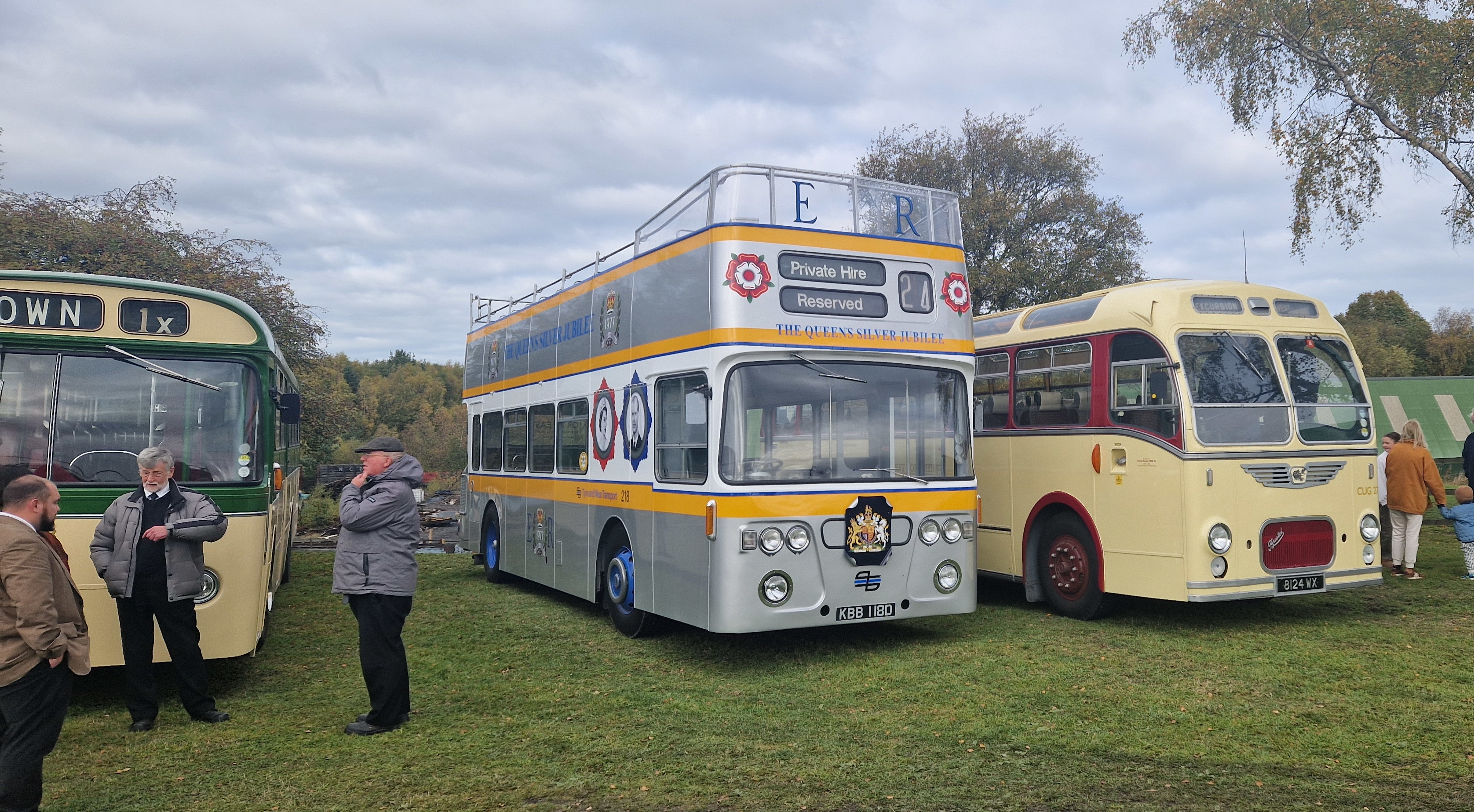 Three vintage buses are parked side-by-side outdoors on a grassy area. On the left is a green and cream single-decker bus, in the center is a silver double-decker open-top bus with yellow stripes commemorating the Queen's Silver Jubilee, and on the right is a cream and red vintage single-decker coach. Several people are standing nearby, viewing the vehicles on an overcast day.