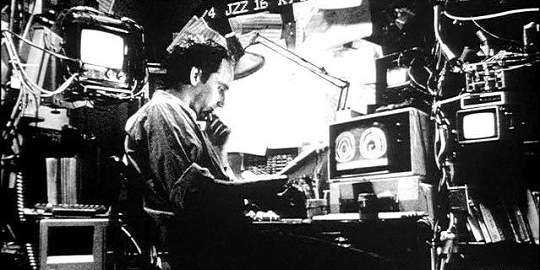 Black and white film still of a man in thought surrounded by grimy computer wiring