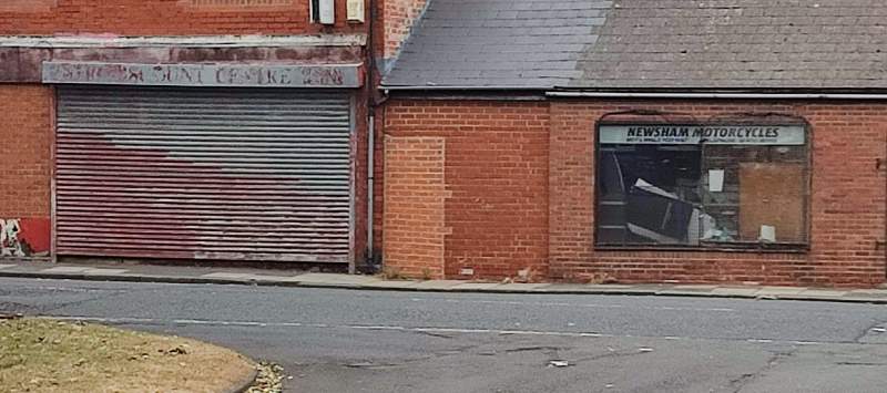 Two empty, shuttered storefronts. One's text is too faded to read, but the other reads 'Newsham Motorcycles'.