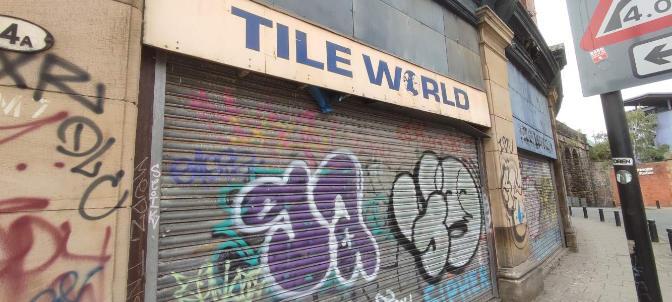 The frontage of a shop by the name of “Tile World” (with a globe replacing the O), its shutters now covered with graffiti.