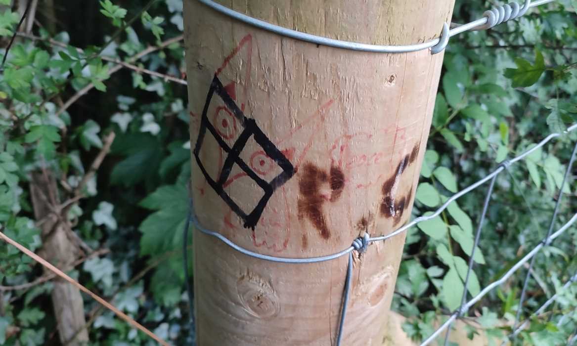 A fencepost crudely vandalised with some sort of four-way grid, an owl saying “Peace”, and the burnt-in initials of one “R.C.”